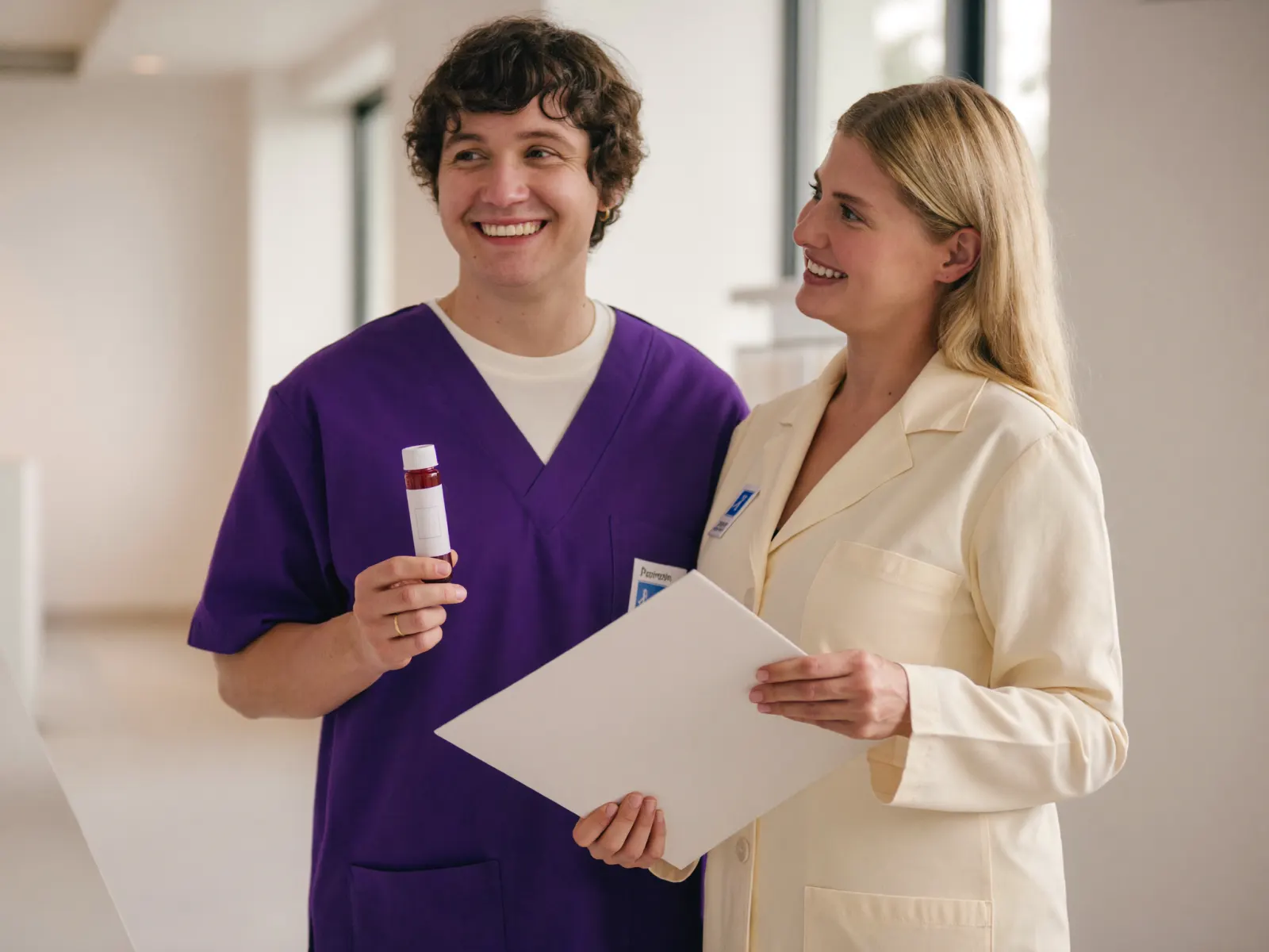 Two smiling clinicians reviewing a labeled blood sample vial and a results folder in a bright clinic